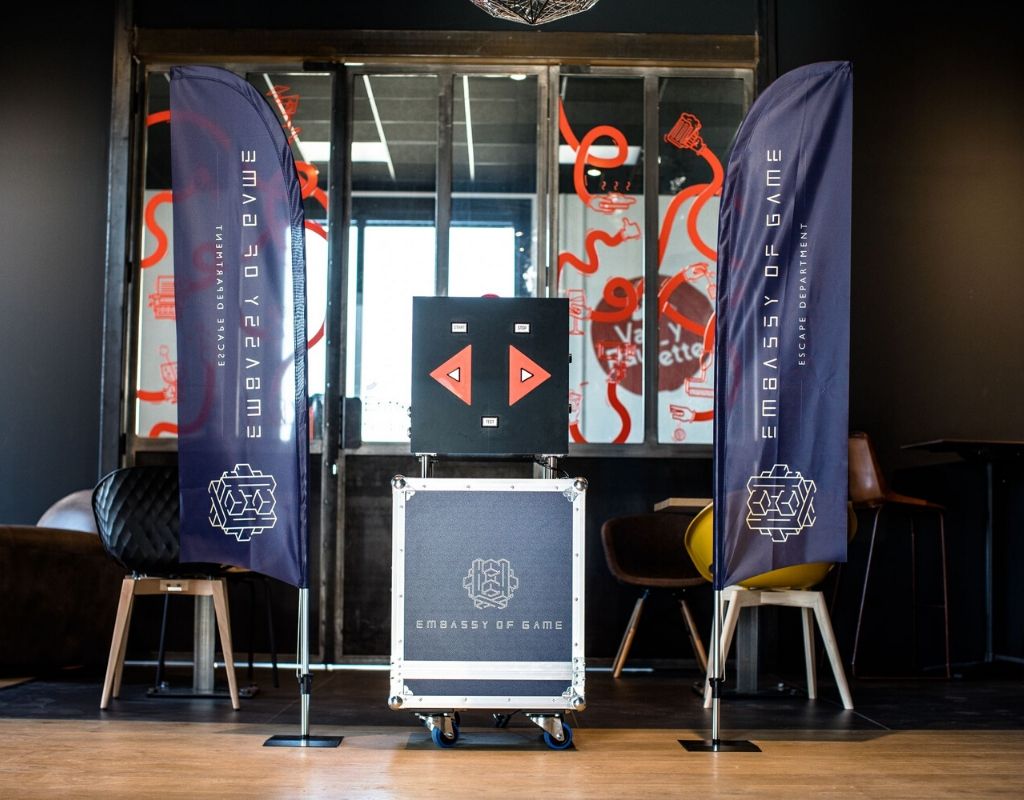 OMNI escape game mobile est posé sur une caisse de transport gravée à l'effigie d'embassy of game de chaque côté des oriflammes bleu foncé