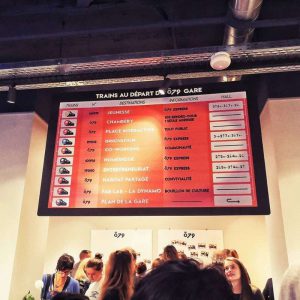 des visiteurs venus inaugurés le lieu tentent de déchiffrer un panneau accroché dessus sont représentées des lignes de train fictives dédiées au jeu