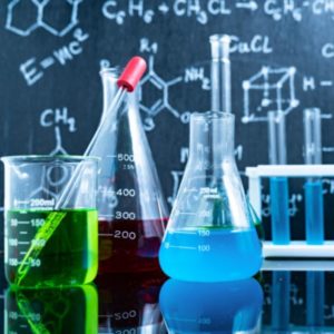 devant un tableau noir où sont inscrits des formules scientifiques à la craie, des béchers et récipients sont remplis de produits colorés rouge, vert et bleu